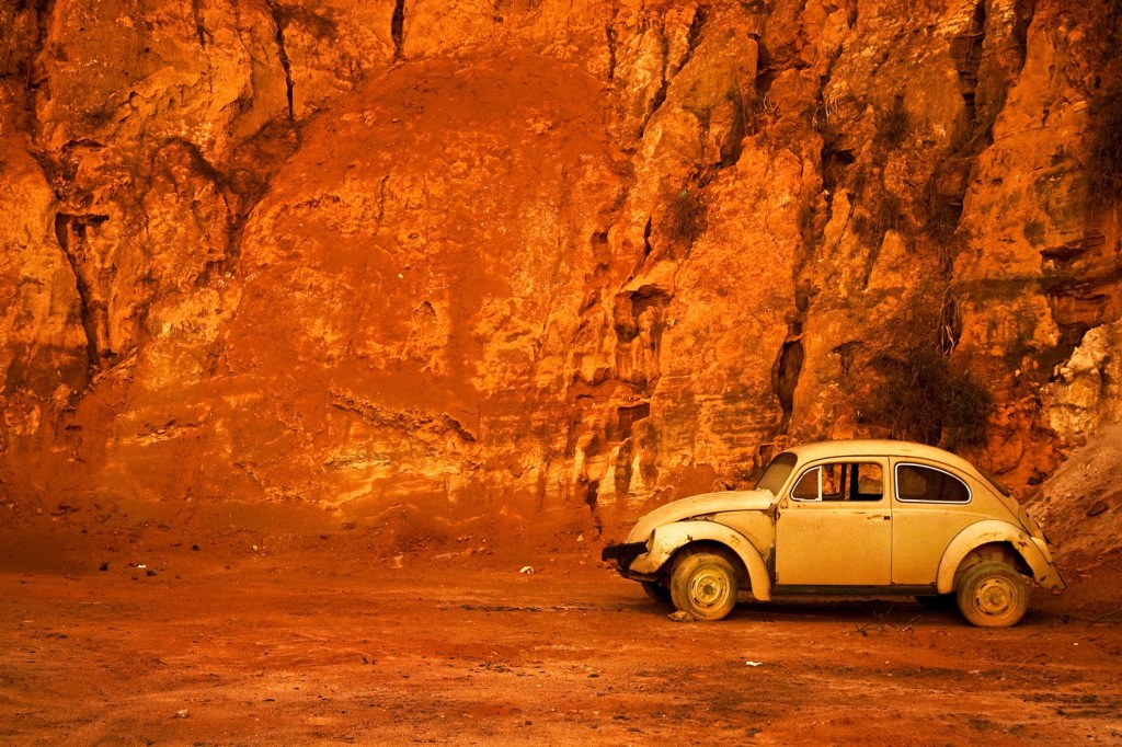 A photograph of an old yellow Volkswagen Beetle car in front of a cliff side of red dirt.